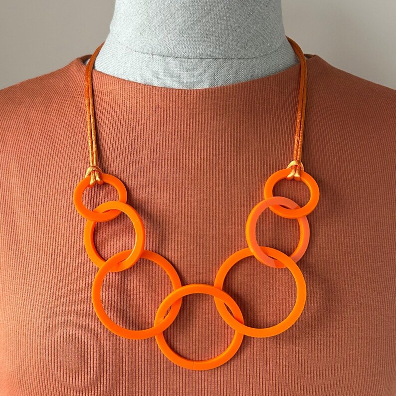 May include: An orange necklace featuring multiple interlocking circular links. The necklace is suspended from a thin, matching orange cord. The design is modern and bold, with a focus on geometric shapes and vibrant color.