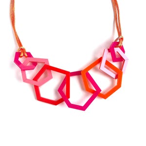 May include: A necklace featuring a series of interlocking hexagonal shapes in shades of bright pink, orange, and light pink. The geometric pendants are suspended from a thin, textured orange cord that connects to the outer hexagons on each side.