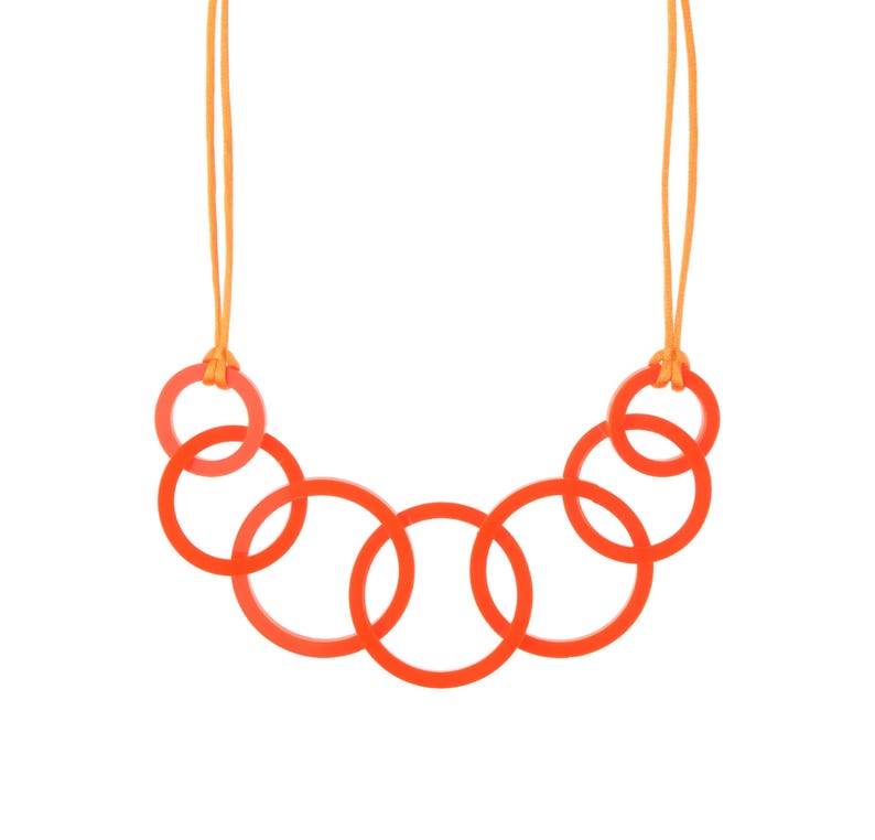 May include: A vibrant orange necklace featuring a series of interconnected circular links. The necklace is suspended by a thin, orange cord. The design is modern and minimalist, with a focus on geometric shapes and bold color.