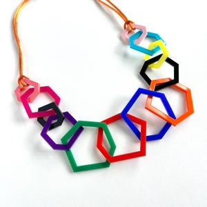 May include: A necklace featuring a series of interlocking, multi-coloured geometric hexagon shapes made of smooth plastic. The vibrant colours include red, orange, yellow, green, blue, purple, pink, and black, all suspended from a thin orange cord.