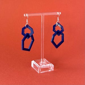 Peut inclure: Une paire de boucles d'oreilles pendantes bleu foncé composées de deux formes hexagonales entrelacées suspendues à des crochets en métal argenté. Les boucles d'oreilles sont présentées sur un support en acrylique transparent en forme de T.