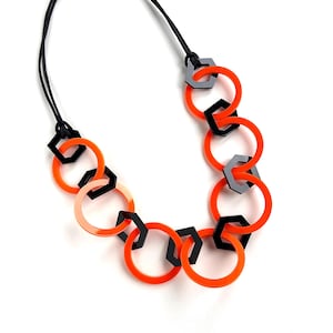 May include: A necklace featuring a series of interlocking bright orange circular rings and smaller black hexagonal shapes. The geometric components are connected in a continuous chain pattern and suspended from a thin black cord for a structured look.