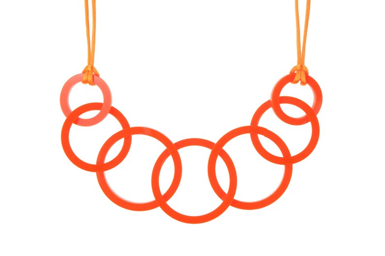 May include: A necklace featuring a series of interlocking orange rings. The necklace is suspended from an orange cord. The rings are a vibrant orange color, and the cord is a lighter orange. The necklace is a modern and minimalist design.