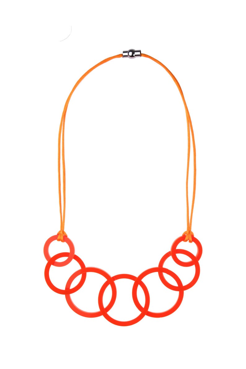 May include: A modern necklace featuring six vibrant red circular pendants connected by an orange cord. The cord is secured with a silver-colored magnetic clasp. The necklace is a bold statement piece.
