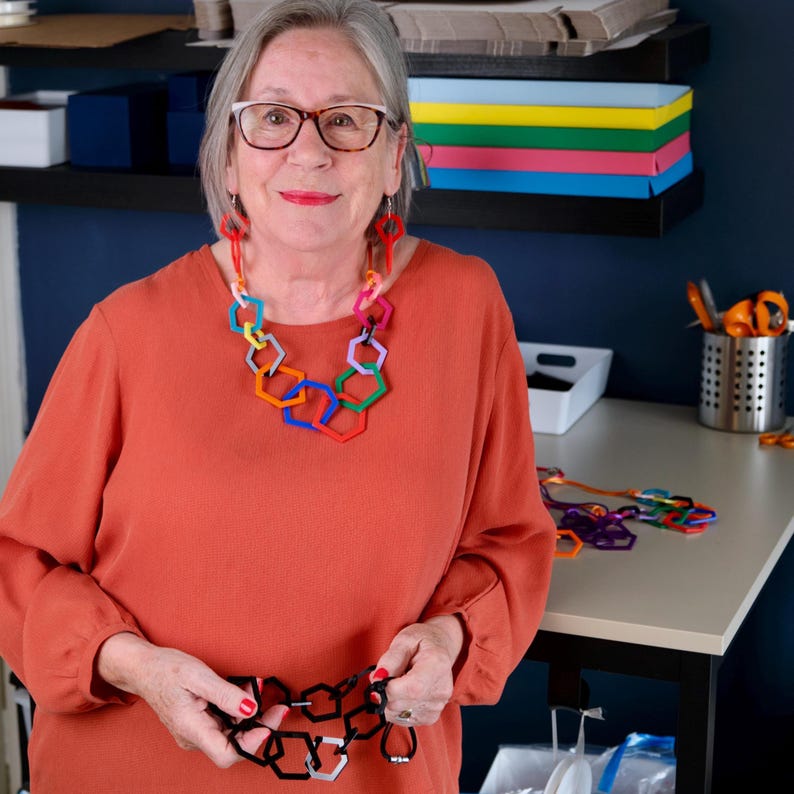 May include: A woman wearing an orange blouse and eyeglasses, with a colorful geometric necklace. The necklace features interlocking hexagonal shapes in various colors. She is holding a black and silver geometric necklace.