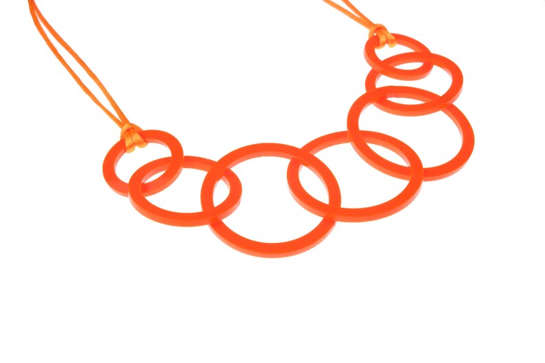 May include: An orange necklace featuring a series of interconnected circular links. The necklace is suspended from a thin orange cord. The design is modern and minimalist, with a focus on geometric shapes and bright color.