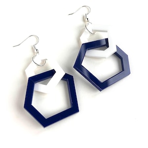 May include: A pair of dangle earrings featuring interlocking geometric shapes. Each earring consists of a smaller white hexagon linked to a larger navy blue hexagon, suspended from silver-toned metal fishhook ear wires for pierced ears.