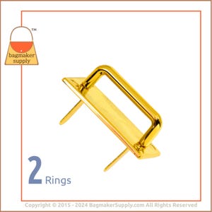 Puede incluir: Dos anillos de metal dorado con puntas para sujetar a una bolsa u otro artículo. Los anillos son rectangulares con una parte superior redondeada y un acabado liso.