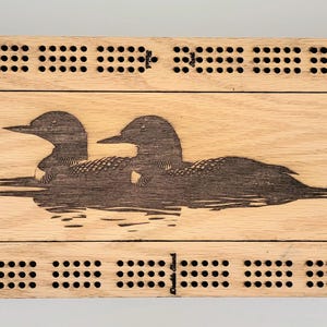 May include: A wooden cribbage board with a laser-etched design of two loons swimming on water. The board is rectangular with rounded corners and features a track of holes for pegs. The loon design is dark brown.