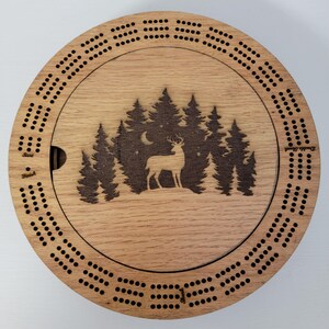May include: A round wooden cribbage board with a forest and deer design. The board has a lid with a laser-etched image of a deer in front of a forest and moon. The board has holes for pegs and the words "Start" and "Finish".