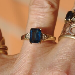 Pu&ograve; includere: Un anello d'oro con una grande pietra preziosa blu scuro di taglio smeraldo circondata da due piccole pietre preziose chiare.