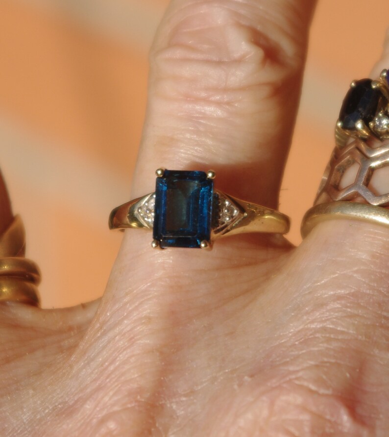 Pu&ograve; includere: Un anello d'oro con una grande pietra preziosa blu scuro di taglio smeraldo circondata da due piccole pietre preziose chiare.