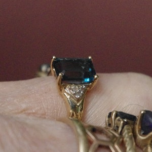 Pu&ograve; includere: Un anello d'oro con una grande pietra preziosa blu scuro a taglio smeraldo circondata da piccole pietre preziose trasparenti.