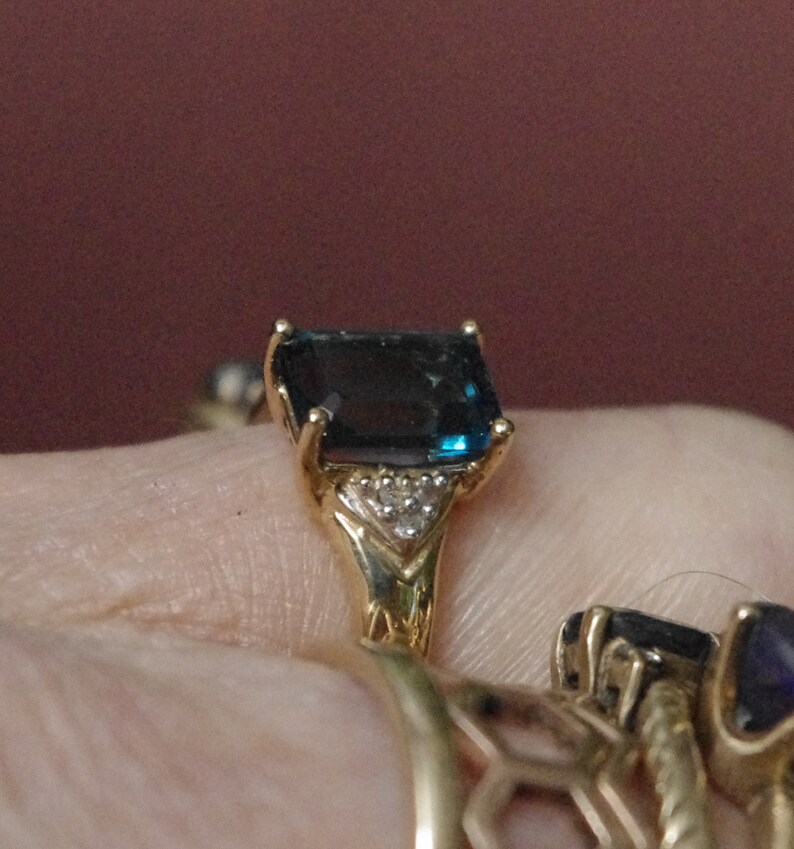 Pu&ograve; includere: Un anello d'oro con una grande pietra preziosa blu scuro a taglio smeraldo circondata da piccole pietre preziose trasparenti.