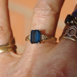 Pu&ograve; includere: Un anello d'oro con una grande pietra preziosa rettangolare blu scuro circondata da pietre preziose pi&ugrave; piccole e trasparenti.