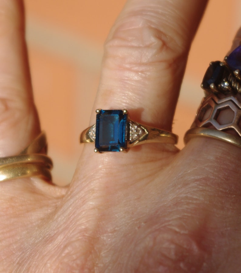 Pu&ograve; includere: Un anello d'oro con una grande pietra preziosa rettangolare blu scuro circondata da pietre preziose pi&ugrave; piccole e trasparenti.