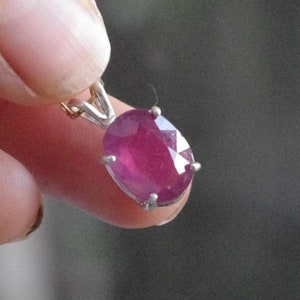 May include: A silver pendant featuring a large oval-shaped ruby gemstone. The ruby is a deep pink colour and has a faceted cut.