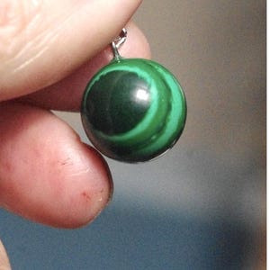 Puede incluir: Un colgante de piedra de malaquita verde con una anilla de plata. La malaquita tiene un patrón de remolino de verde oscuro y claro.