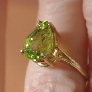 May include: A gold ring featuring a pear-shaped, light green gemstone. The gemstone is faceted, catching the light and displaying a vibrant, lime green color. The ring's band is a simple, elegant design.