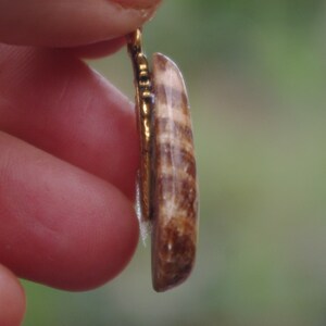 May include: A close-up of a gold pendant with a brown and white striped stone. The pendant is held by a finger.