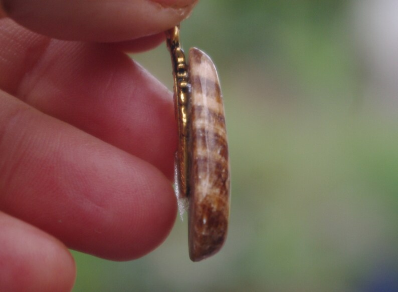 May include: A close-up of a gold pendant with a brown and white striped stone. The pendant is held by a finger.