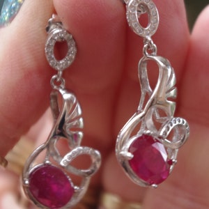 May include: A pair of silver earrings featuring a ruby gemstone and a delicate, intricate design. The earrings have a small circle of clear stones at the top and a larger ruby gemstone at the bottom.