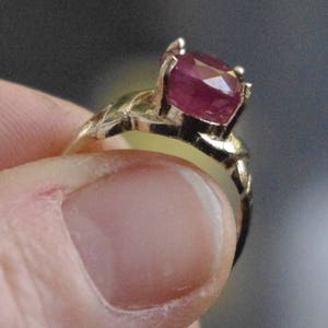 May include: A gold ring featuring a large, faceted ruby gemstone. The ruby is a deep red color and is set in a four-prong setting. The ring's band has an intricate, textured design.