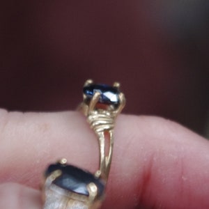 May include: A gold ring with two dark blue oval-shaped gemstones. The ring is on a finger.
