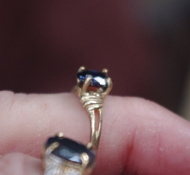 May include: A gold ring with two dark blue oval-shaped gemstones. The ring is on a finger.