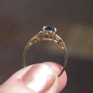 Puede incluir: Un anillo de oro con una piedra preciosa morada facetada de forma ovalada, engastada en una montura de cuatro garras. El aro presenta un dise&ntilde;o fino y calado con acabado met&aacute;lico, mostrando la vista lateral del engaste y el tono profundo.