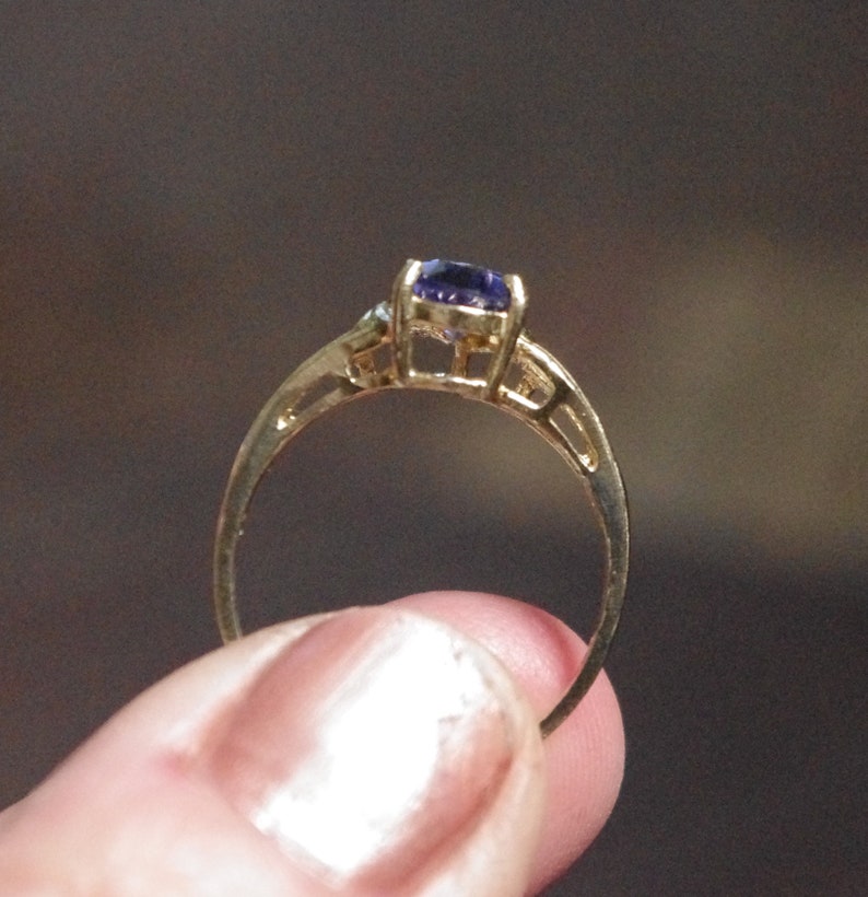 Puede incluir: Un anillo de oro con una piedra preciosa morada facetada de forma ovalada, engastada en una montura de cuatro garras. El aro presenta un dise&ntilde;o fino y calado con acabado met&aacute;lico, mostrando la vista lateral del engaste y el tono profundo.