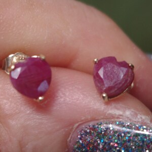 Peut inclure: Une paire de boucles d'oreilles en rubis en forme de cœur avec des montures en or. Les boucles d'oreilles sont présentées sur la main d'une personne.