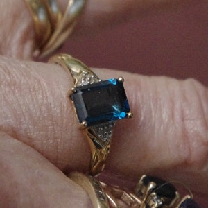 Pu&ograve; includere: Un anello d'oro con una grande pietra preziosa blu scuro a taglio smeraldo circondata da pietre preziose pi&ugrave; piccole e trasparenti.
