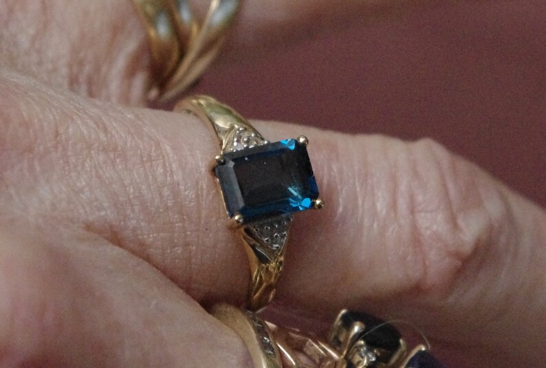 Pu&ograve; includere: Un anello d'oro con una grande pietra preziosa blu scuro a taglio smeraldo circondata da pietre preziose pi&ugrave; piccole e trasparenti.