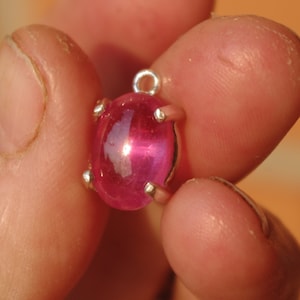 May include: A polished, oval pink gemstone pendant with a silver metal setting. The gemstone is held by four small prongs. A small loop at the top is for attaching to a chain or bracelet. The pendant is held between fingers.