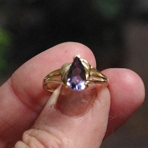 Puede incluir: Un anillo de oro con una piedra preciosa morada en forma de pera en el centro. La banda del anillo tiene un dise&ntilde;o dividido en ambos lados, que conduce al engaste de garras que sujeta firmemente la piedra violeta en forma de l&aacute;grima.