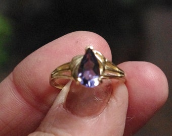 May include: A gold ring featuring a pear-shaped purple gemstone set in the center. The band of the ring has a split design on both sides, leading up to the prong setting that holds the teardrop-shaped violet stone securely in place.