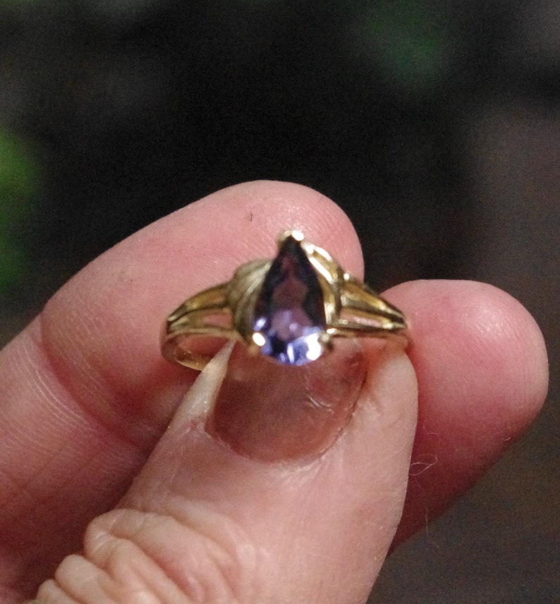 Puede incluir: Un anillo de oro con una piedra preciosa morada en forma de pera en el centro. La banda del anillo tiene un dise&ntilde;o dividido en ambos lados, que conduce al engaste de garras que sujeta firmemente la piedra violeta en forma de l&aacute;grima.