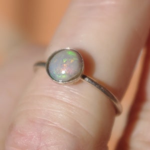 May include: A silver ring with a round opal gemstone. The opal has a white base with iridescent flashes of green, blue, and pink.