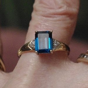 Pu&ograve; includere: Un anello d'oro con una grande pietra preziosa rettangolare blu circondata da pietre preziose pi&ugrave; piccole e trasparenti.