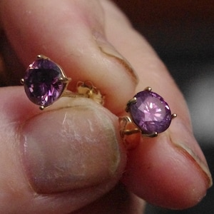 Könnte beinhalten: Ein Paar goldene Ohrringe mit violetten Edelsteinen. Die Ohrringe werden in der Hand einer Person gehalten.