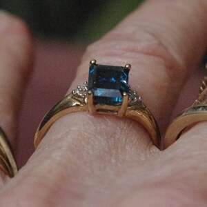 Pu&ograve; includere: Un anello d'oro con una grande pietra preziosa di colore blu scuro, di taglio smeraldo, circondata da pietre preziose pi&ugrave; piccole e trasparenti.