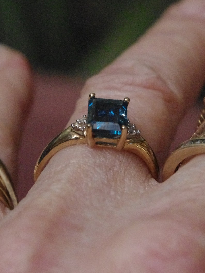 Pu&ograve; includere: Un anello d'oro con una grande pietra preziosa di colore blu scuro, di taglio smeraldo, circondata da pietre preziose pi&ugrave; piccole e trasparenti.