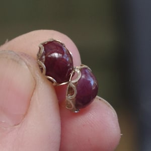 May include: A pair of small, round earrings with a deep burgundy, glossy finish. The earrings are set in a silver-coloured metal with an open, filigree design. The earrings are held in a person's hand.