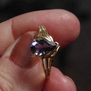 Puede incluir: Un anillo de oro con una piedra preciosa morada en forma de pera montada en un engaste de garras. La banda del anillo tiene un dise&ntilde;o dividido que se estrecha hacia el centro, creando una apariencia delicada y estructurada alrededor de la piedra.