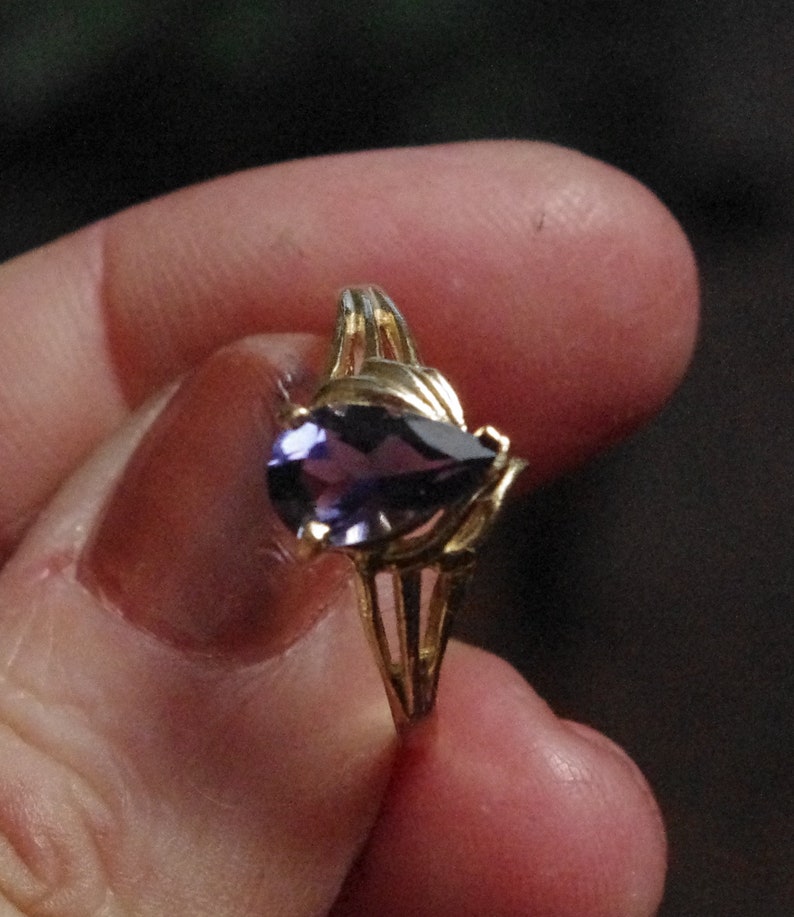 Puede incluir: Un anillo de oro con una piedra preciosa morada en forma de pera montada en un engaste de garras. La banda del anillo tiene un dise&ntilde;o dividido que se estrecha hacia el centro, creando una apariencia delicada y estructurada alrededor de la piedra.