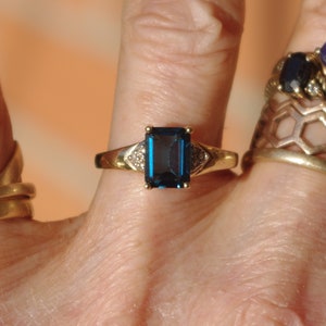 Pu&ograve; includere: Un anello d'oro con una grande pietra preziosa blu scuro a taglio smeraldo circondata da pietre preziose pi&ugrave; piccole e trasparenti.