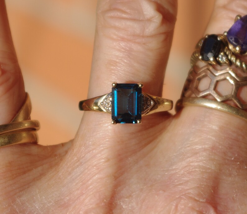 Pu&ograve; includere: Un anello d'oro con una grande pietra preziosa blu scuro a taglio smeraldo circondata da pietre preziose pi&ugrave; piccole e trasparenti.