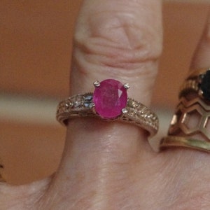 Op de afbeelding: Een zilveren ring met een grote ovale roze edelsteen, omringd door kleinere heldere edelstenen.