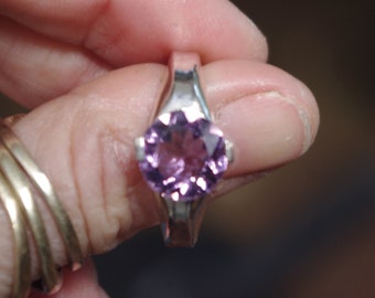 May include: A silver ring featuring a light purple gemstone. The ring has a simple design with a smooth band and a round gemstone.